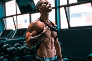 Muscular man showing abs and tattoos in a gym setting.